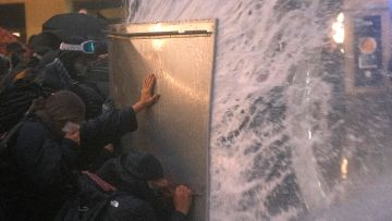 Demonstrant:innen schützen sich mit einem Schild vor dem Strahl des Wasserwerfers