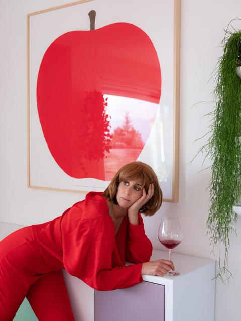 Selbstportrait-Inszenierung von Nora Rupp: Sie steht vor einem Gemälde eines Apfels und schaut mit gedrehtem Kopf ins Leere, daneben steht ein Glas Wein