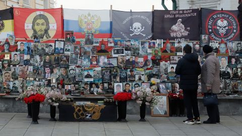 eine Wand mit Porträts von Gefallenen unter den Flaggen der «russischen Welt», davor stehen zwei Personen und Vasen mit Blumen 