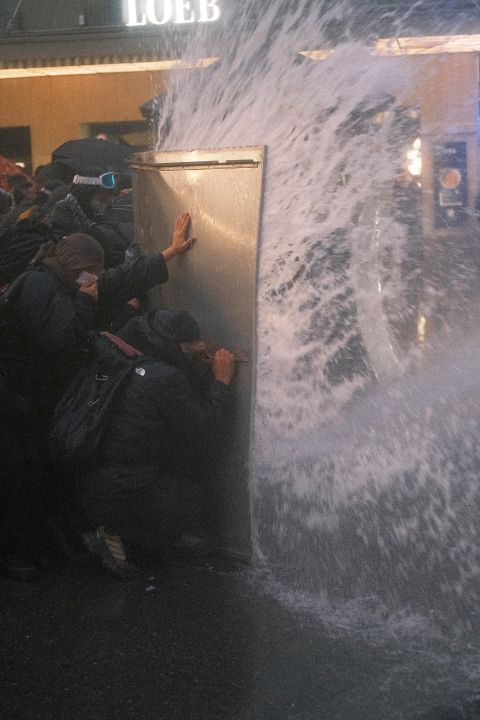 Demonstrant:innen schützen sich mit einem Schild vor dem Strahl des Wasserwerfers
