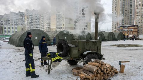 Rettungskräfte stehen vor einem mobilen Holzofen, welcher ein städtisches Notwärmezentrum in Kyjiw beheizt