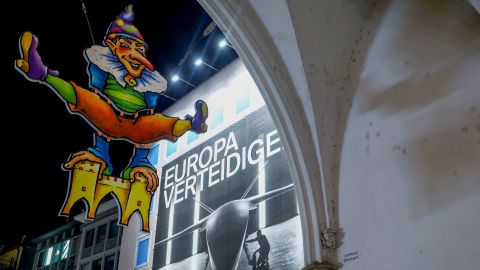 Plakat der deutschen Firma Hensoldt mit Abbildung einer Kampfdrohne beim Karlstor in München, im Vordergrund ein Schild mit Fasnacht-Sujet