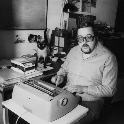 Karl Lüönd sitzt an der Schreibmaschine, daneben steht seine Katze auf einem Tisch