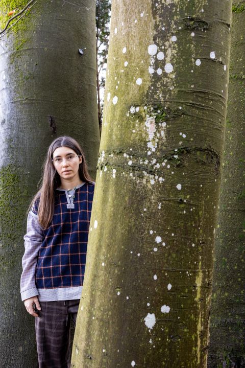 Clara Heinrich schaut hinter einem Baum im Wald hervor