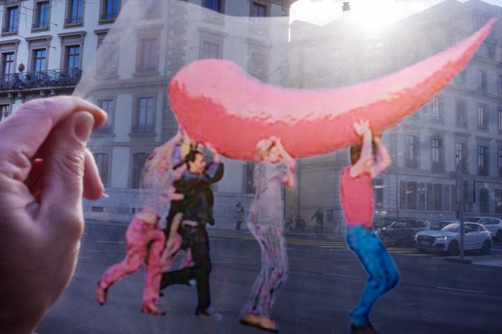 Fotografie einer Folie, auf der Menschen zu sehen sind, welche ein übergrosses rotes Nashorn-Horn tragen, im Hintergrund ein Gebäude in Genf