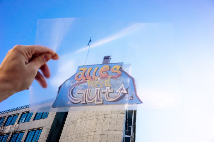 Fotografie einer Folie, auf der das Wohlgroth-Graffiti «Alles wird Gut» zu sehen ist, im Hintergrund ein modernes Gebäude