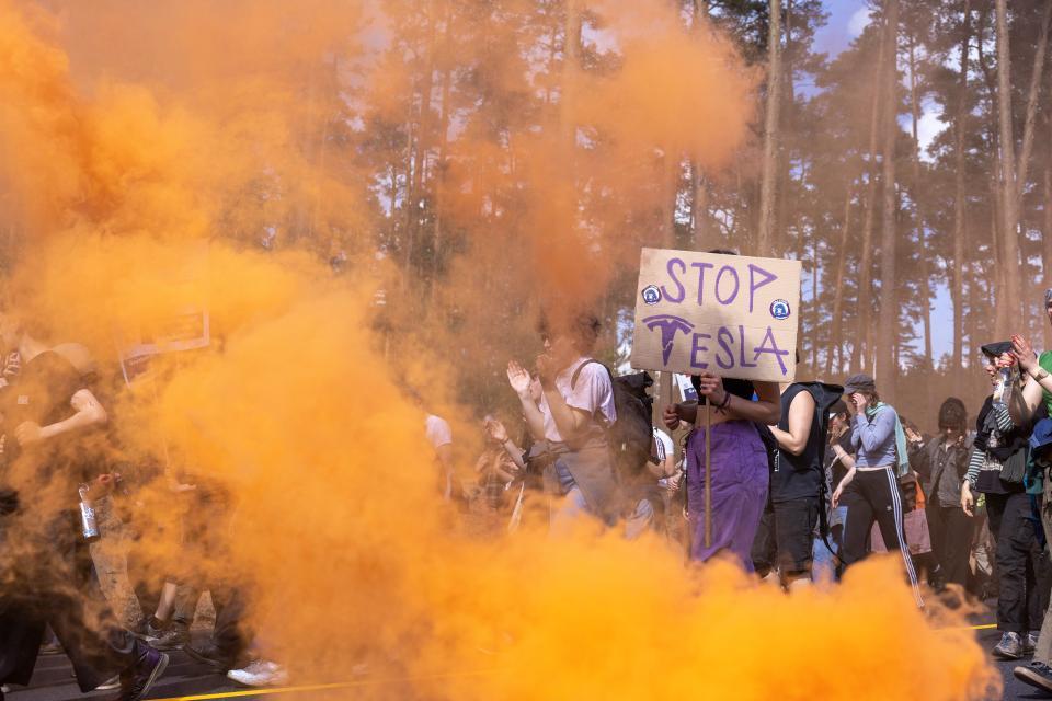 Protestbild aus einem Wald mit orangem Rauch und einem Schild, auf dem es «Stop Tesla» heisst.