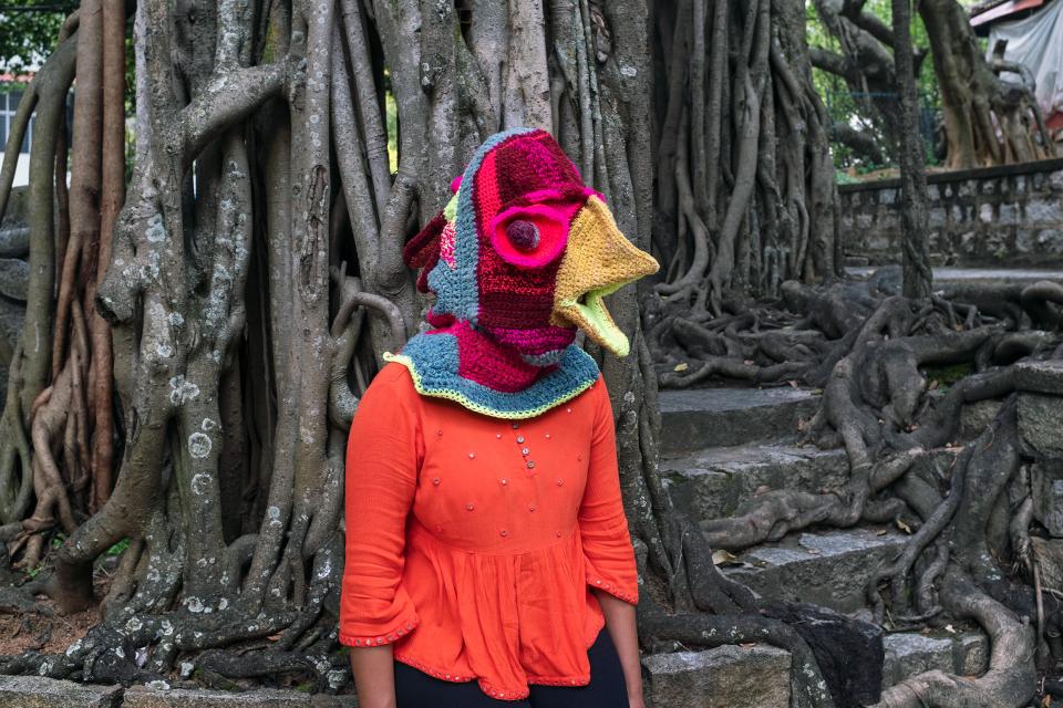 Fotoinszenierung: eine Person mit einer Mütze in Vogelform, welche den Kopf verdeckt, steht vor einem Baum welcher Treppen und Mauern überwuchert.
