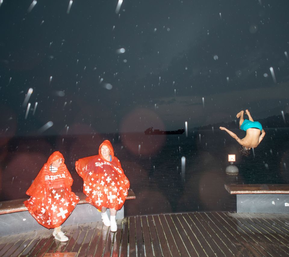 zwei ältere Frauen sitzen in Regenpelerinen am See, daneben springt ein junger Mann im Salto ins Wasser