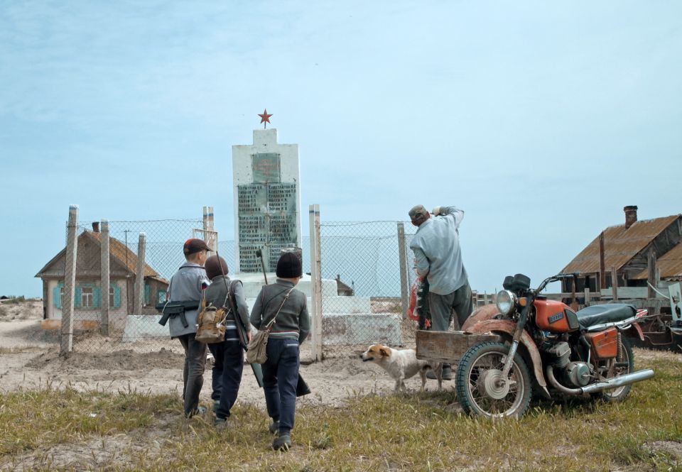 Filmstill aus «Ostrov»: drei Kinder und ein Mann mit einem Motorad vor einem Denkmal