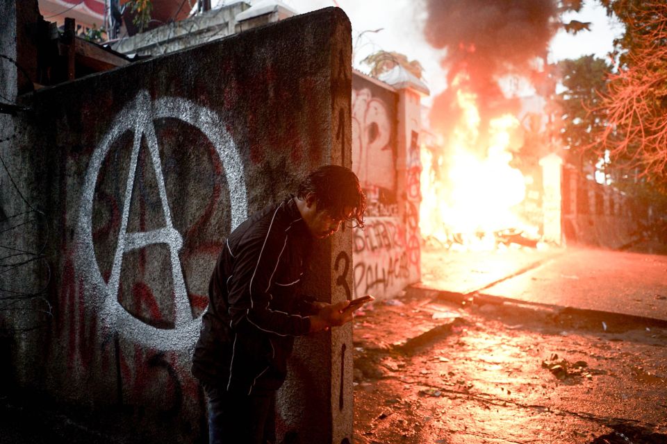 eine Person schaut vor einem brennenden Regierungsgebäude auf ihr Smartphone