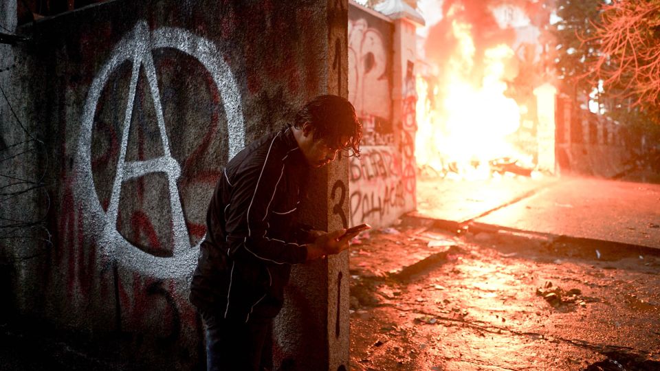 eine Person schaut vor einem brennenden Regierungsgebäude auf ihr Smartphone