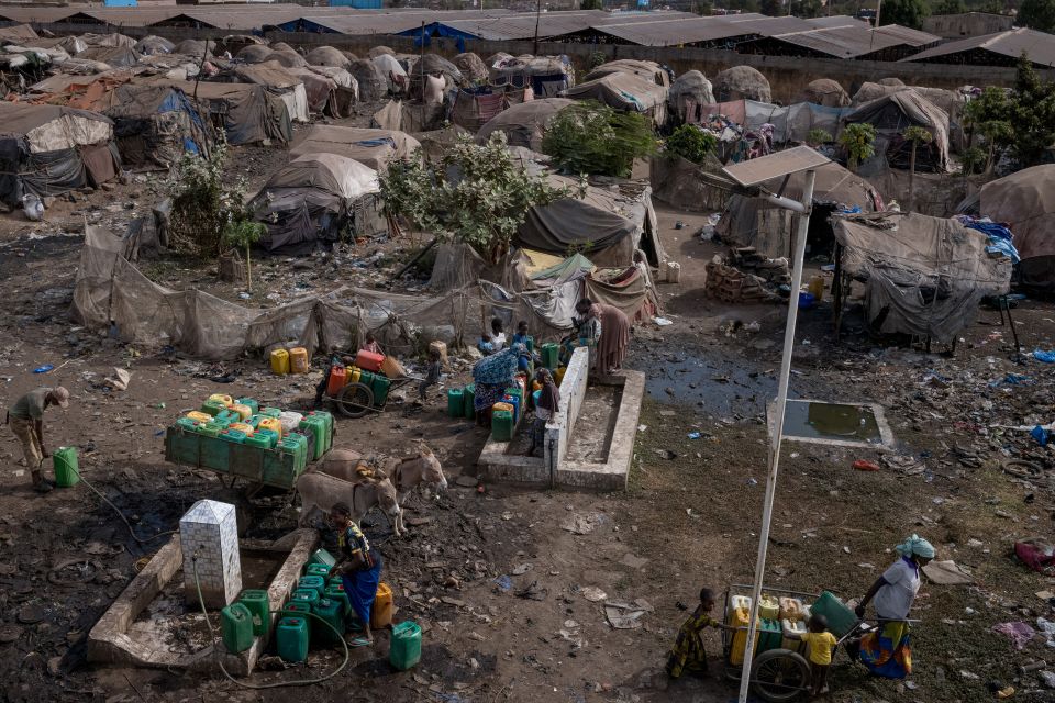 Menschen füllen Wasser an einem Wasserverteiler ab, im Hintergrund improvisierte Zelt-Behausungen