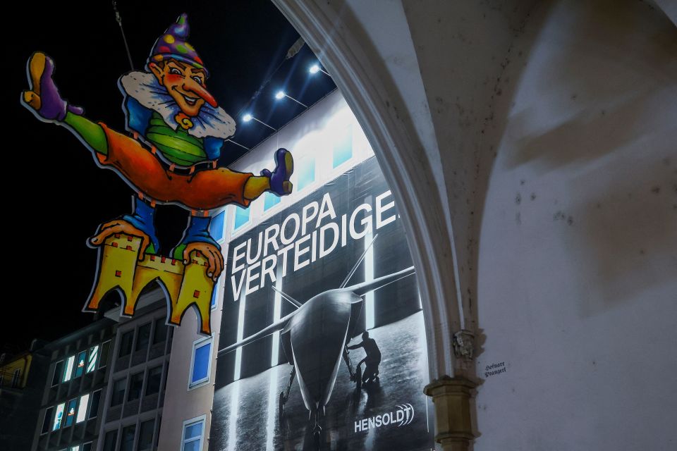 Plakat der deutschen Firma Hensoldt mit Abbildung einer Kampfdrohne beim Karlstor in München, im Vordergrund ein Schild mit Fasnacht-Sujet