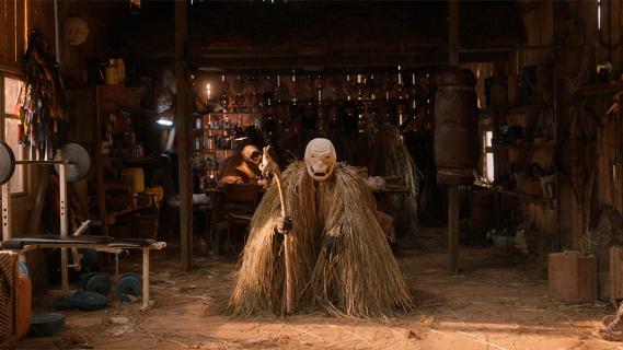 Filmstill aus «Pamfir»: eine unheimliche Figur mit Holz-Maske und Strohüberwurf steht in einem Schupen 