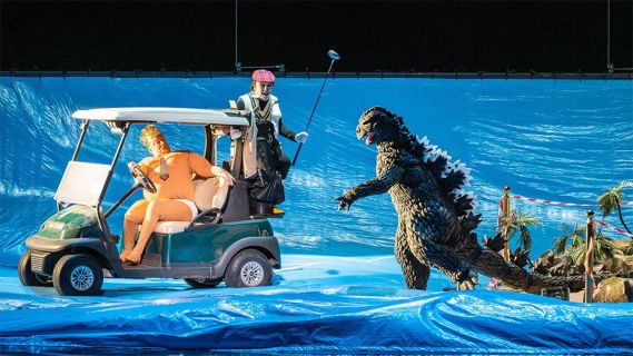 Bühnenfoto der Oper «Monster’s Paradise»: zwei Personen auf einem Golfmobil, daneben eine Godzilla-Figur